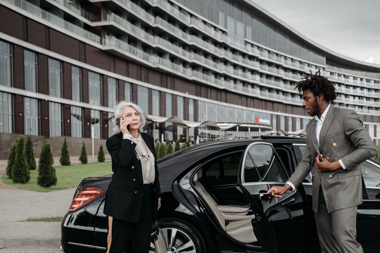 Crafting Captivating Headlines: Your awesome post title goes here Senior female executive on phone with chauffeur holding car door open, modern architecture backdrop.