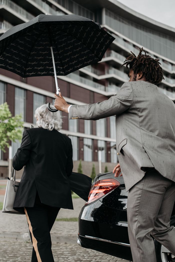 our-story Two business professionals with an umbrella near a luxury car in an urban setting.