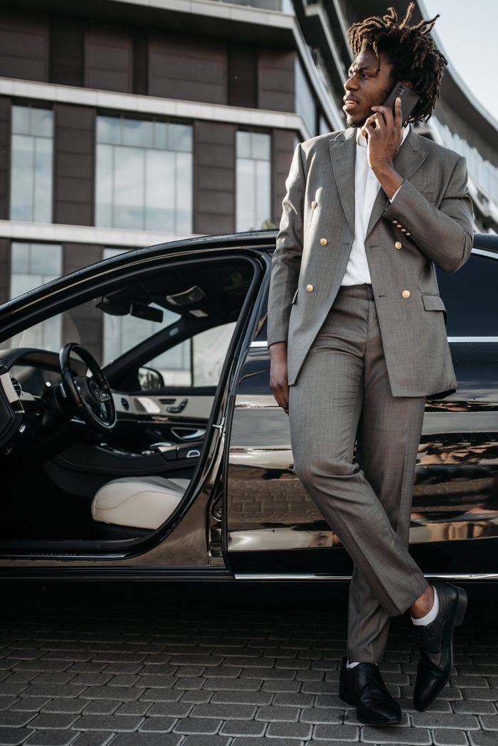 about-us Stylish businessman in suit using mobile phone while leaning against a black luxury car.
